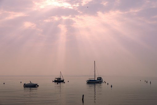 The evening sun rays pierce the clouds, casting a soft, ethereal glow over the boats and waterfowl on Lake Garda, all enveloped in a mist of gentle rose hues