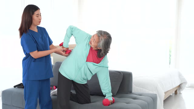 Concept of care and support. A woman in a blue uniform is helping an older woman lift weights on a couch.