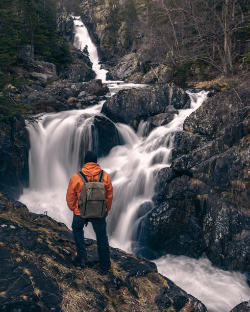istock A man in an orange jacket is standing on a rock overlooking a waterfall. 2149492716