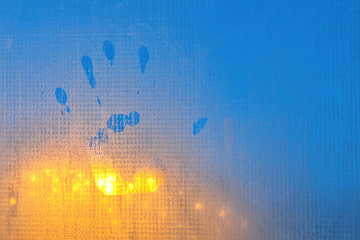 A handprint on the foggy surface of a plastic window. In the background there is the blue of the evening sky and the yellow of the street lights. Background.