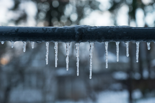 Several icicles of different lengths hang from the gray pipe of the crossbar. An evening blurry landscape with trees is visible in the background. Background.