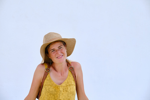 Positive adult female tourist wearing hat and casual clothes looking away while standing against white wall on sunny summer day
