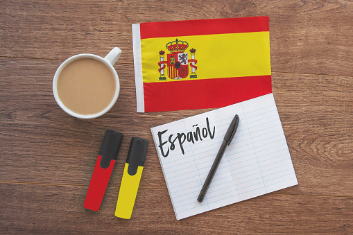spain national flag, notebook with the inscription spanish in spanish, two markers, cup of coffee on brown wooden desktop, flat way, concept of learning foreign languages