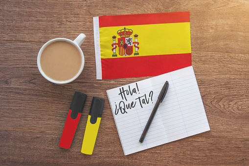 spain national flag, notebook with the inscription hello, how are you in spanish, two markers, cup of coffee on brown wooden desktop, study concept 1