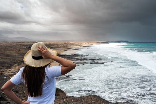 A woman wearing a straw hat stands on a rocky beach looking out at the ocean. The sky is cloudy and the water is choppy