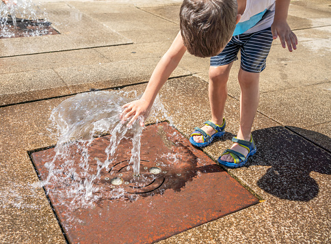 A child play in the square with the water from the fountain on a sunny summer day.Active summer leisure for kid in the city.