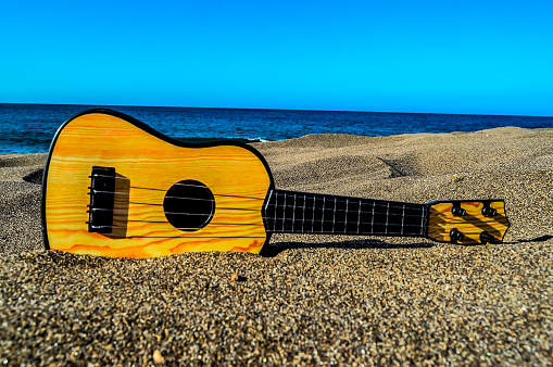 A small guitar is laying on the sand next to the ocean. The guitar is yellow and black