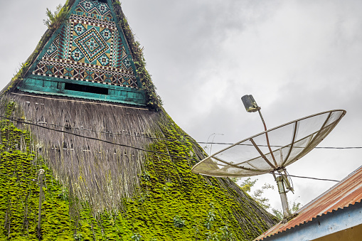 Merek, North Sumatra, Indonesia - January 28th 2024:  House with a classical Batak roof and a dish antenna. The thatched roof is covered in moss but the gable has the traditional pattern decoration