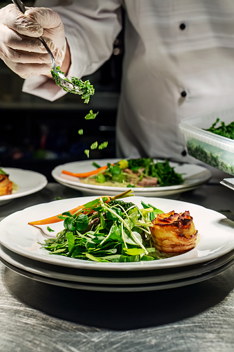 Chef working at the pass adds dill-flavoured flakes to a dish of ballotine of veal mixed with fresh wild garlic leaves or ramsons and served seasonal vegetables with a spinach-flavoured sauce.