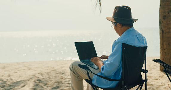 Asian elderly man sitting on a beach chair enjoys the sea view. A happy senior adult man enjoys the travel lifestyle after retirement concept.