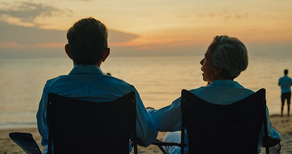 Asian elderly couple holding each other hand while sitting on a beach chair enjoys the sea view at sunset light. A happy senior adult people enjoy the travel lifestyle after retirement concept.