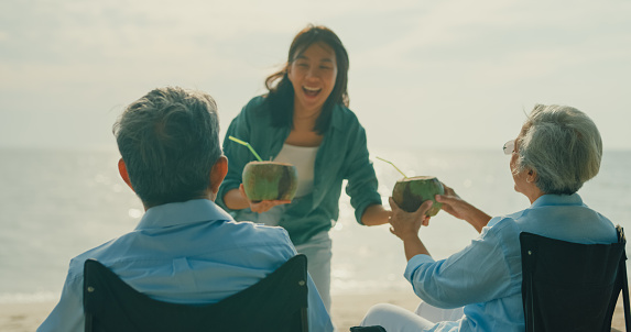 Asian elderly cheerful couple drinking coconut while sitting on a beach chair enjoying the sea view. A happy senior adult people enjoy the travel lifestyle after retirement concept.