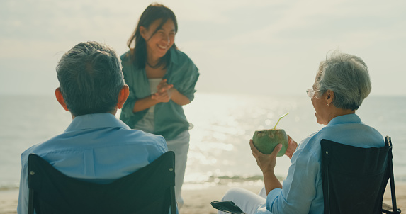 Asian elderly cheerful couple drinking coconut while sitting on a beach chair enjoying the sea view. A happy senior adult people enjoy the travel lifestyle after retirement concept.