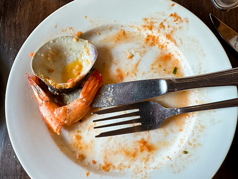 The remains of a meal featuring seafood are strewn across a white plate. An empty shell, a solitary shrimp tail, and smudges of sauce