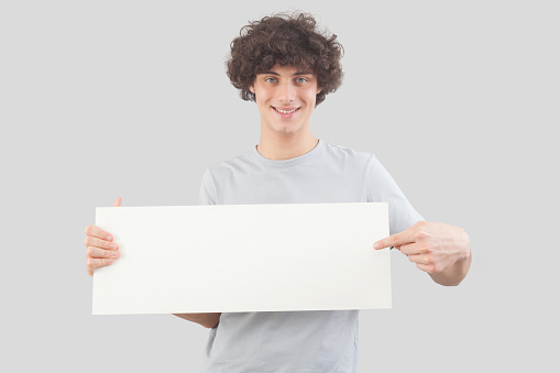 Young man, smiling and handsome, pointing with finger to show a blank white signboard, isolated on gray background. Placard copy space for text or logo, banner for advertising or promotional messages