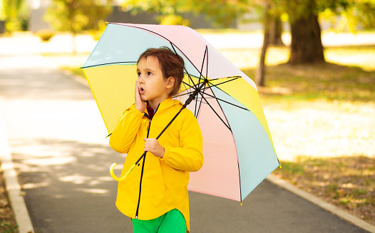 Little cute girl kid child in a yellow raincoat with multicolored rainbow umbrella looks at the sky
