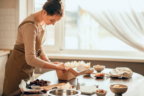 A chef in an apron works in a sunlit kitchen, folding and preparing a box filled with pastries and other delights.