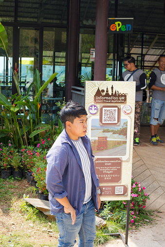 Adult thai man is standing outside of  Pang Kha Royal Project And Development Centre coffee shop in landscape around Pang Kha in north Thailand with Royal Development Center and coffee plantations in mountains around Phayao. Outside are several informational signs with QR codes. Exact location is 9CHV+935, Pha Chang Noi, Pong District, Phayao 56140, Thailand. Man is wearinfg casual clothing and is standing close to one the information billboards. In background are other men outside of entrance