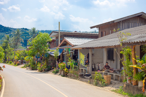 View along road and rural homes in small town in landscape around Pang Kha in north Thailand with Royal Development Center and coffee plantations in mountains around Phayao. Area betwen Ban Hae and Pha Changf Noi  close to Pang Kha Royal Project And Development Centre. In background are mountains. A woman is sitting outside of a house and is relaxing barefoot.
