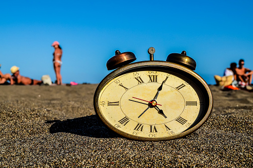 A clock is sitting on the sand with the hands pointing to the number 12. The beach is empty and the sky is blue