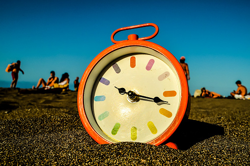 A clock is sitting on the sand with the hands pointing to the number 12. The beach is empty and the sky is blue