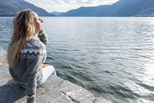 A woman from behind, sitting peacefully at a lake's edge basking in the sun's soft rays. She is dressed in a cozy, patterned sweater and light-colored pants, perfectly blending with the serene environment. She has one hand behind her and the other under her chin. The lake's still waters extend towards the gentle slopes of distant mountains under a soft, overcast sky.