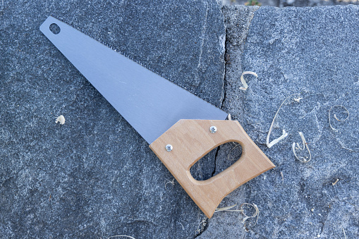 A close-up of a hand saw resting on textured stonework, surrounded by wood shavings. The simplicity of the saw, with its wooden handle and solid grey blade, speaks to manual labor.