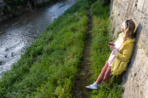 Mature woman pauses on riverbank in springtime and uses mobile phone, Liguria