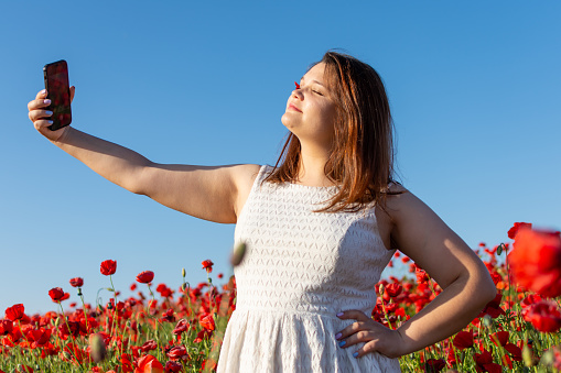 A woman in a white dress smiles while taking a selfie in a field of red flowers under the sunlight. The happy gesture captures the beauty of nature