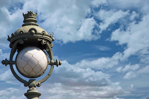 Magnificent cast iron lighting fixture with a white glass shade, installed on a bridge in the city center in the century before last, interesting street lighting technologies, details and decorations, cloudy sky, vintage background, stock photo, summer.