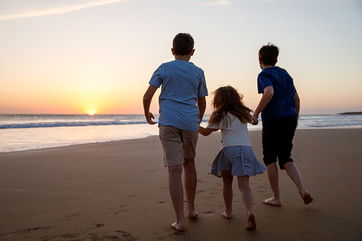 Three kids silhouettes running and jumping on beach at sunset. happy family, two school boys and one little preschool girl. Siblings having fun together. Bonding and family vacation
