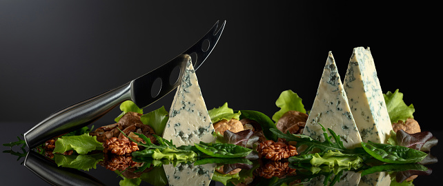 Blue cheese with knife, walnuts and fresh greens on a black background. Copy space.