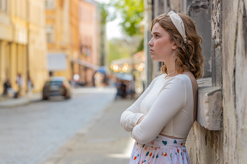 Side view of sad unhappy woman thinks over life concerns, suffers from unfair situation. Problem, feeling bad, annoyed, burnout. Frustrated depressed lonely girl leaning on wall along city street.