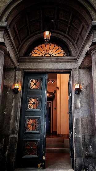 A perspective shot of a big building entrance door with two wings. One side of the door is open and we can see the lobby of the building lit with yellow lighting. The gate is very high with also a haft circular window on the top. There is an arch at the entrance ceiling.