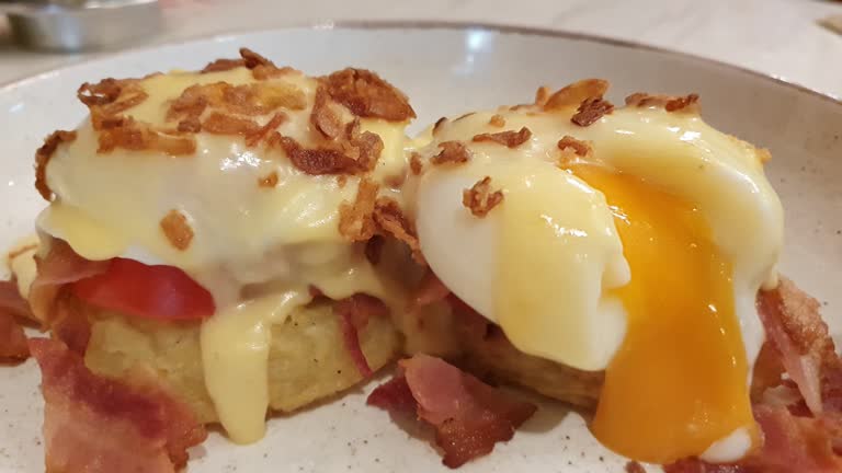 Close up of Eggs Benedict with liquid egg yolk running out. Two poached eggs on potato pancakes or toasted bread with tomatoes, fried onions and bacon with hollandaise sauce