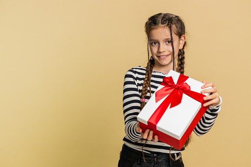 Lovely smiling young preteen child girl kid presenting birthday gift box stretches out hands, offer wrapped present career bonus, celebrating party. Little cute children on studio beige background