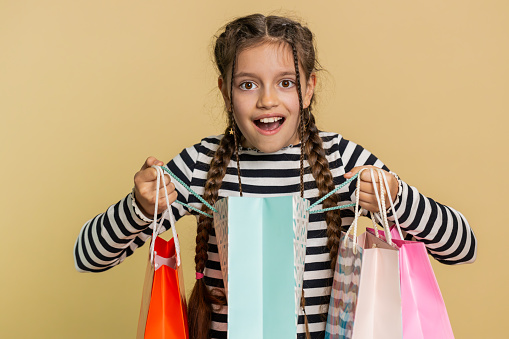 Young preteen child girl kid showing shopping bags, advertising discounts, smiling looking amazed with low prices, shopping on Black Friday holidays. Teenager children isolated on beige background