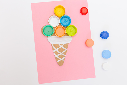 DIY plastic bottle caps for crafts and art, recycling plastic ideas, an ice cream cone craft on a pink background, a pastel, process art, paper, children playing with lids, top view,