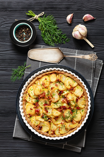 Irish potato pie of crispy crust layered with potatoes, bacon and onion in baking dish on black wooden table with ingredients and cake shovel, vertical view from above