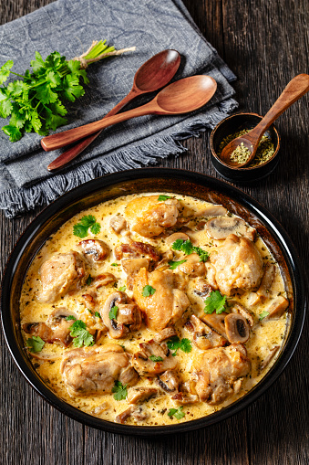 chicken thighs in a creamy mushroom garlic sauce with herbs and parmesan cheese in baking dish on dark wooden table with wooden spoons, vertical view