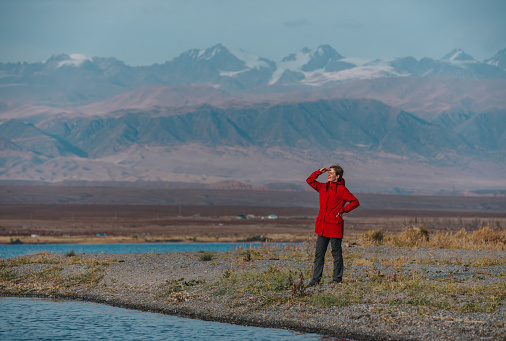 Woman tourist stands on shore of lake on mountains background and looking away