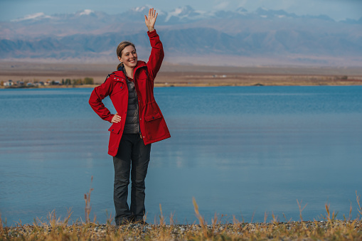 Smiling woman tourist stands on shore of lake on mountains background and waving hand