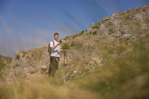 Young handsome hiker using mobile phone, reading a route while resting on the top of a mountain