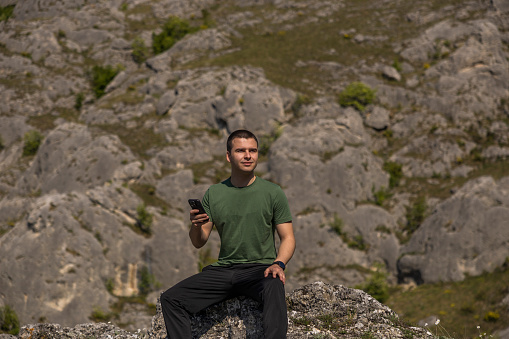 Young handsome hiker using mobile phone while resting on the top of a mountain
