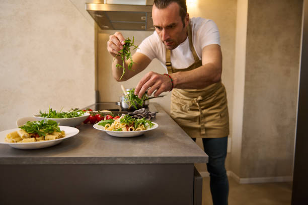 istock Handsome chef cooking delicious Italian pasta with the sauce, seasoning adding fresh arugula leaves and grated parmesan cheese on the pasta in a plate, standing at kitchen counter. Decoration of dish 2149572945