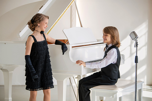 Two children dressed in black and white pose in front of a piano. Scene is playful and lighthearted, as the children are dressed up in formal attire and seem to be enjoying themselves