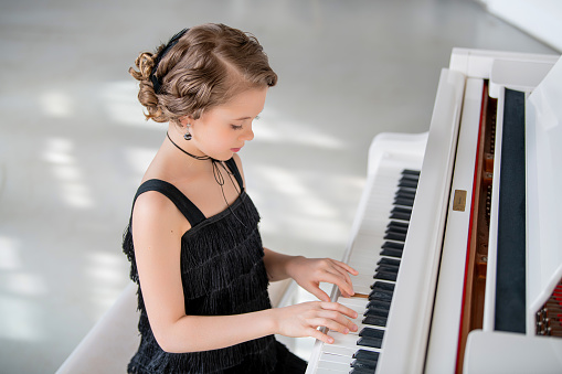 A young girl is playing the piano in a black dress. She is smiling and she is enjoying herself. The piano is white and has a black keyboard