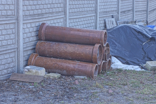 a pile of brown sewer ceramic pipes lie on the ground in green grass on the street near a gray stone fence wall
