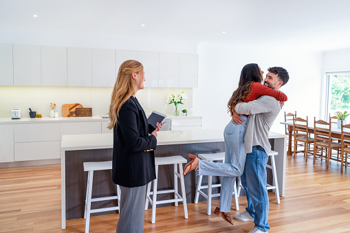 Happy couple embracing and celebrating after buying or renting their new home. The real estate agent is standing beside them with a digital tablet. The kitchen and dining room can be seen in the background