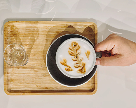 Top view of wooden tray with cup of capuccino latte coffee and glass of water with male hand, foam drink picture, breakfast mood
